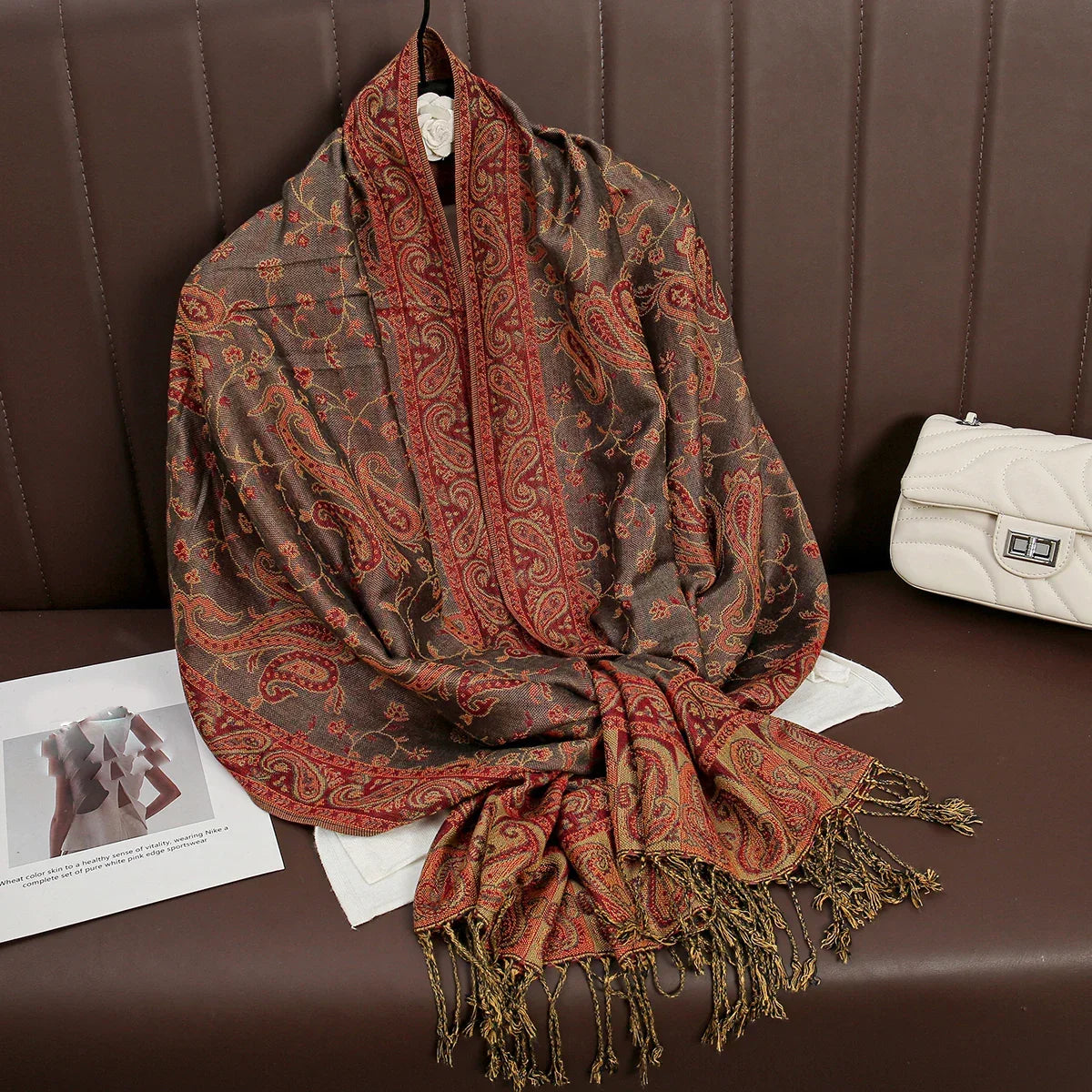 Brown and red paisley pashmina scarf with fringe on brown sofa, white quilted purse beside.