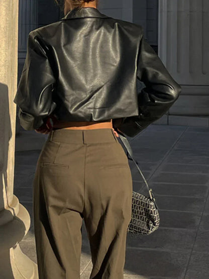 Woman wearing a cropped black leather jacket and high-waisted brown pants outdoors with a designer handbag