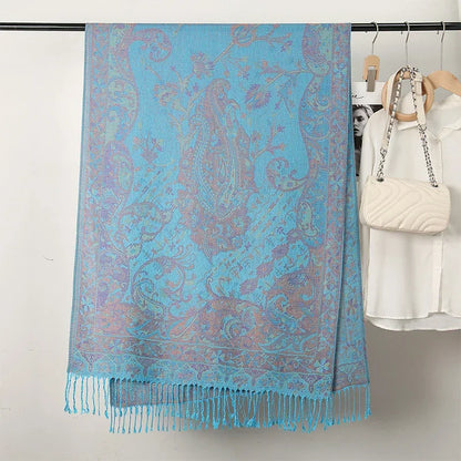 Blue paisley jacquard scarf with fringe, draped on a clothing rack beside a white blouse and handbag