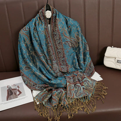 Blue paisley shawl with gold fringe displayed on brown couch next to white purse