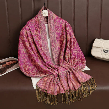 Pink paisley floral shawl with gold fringe draped on a brown couch, white handbag nearby