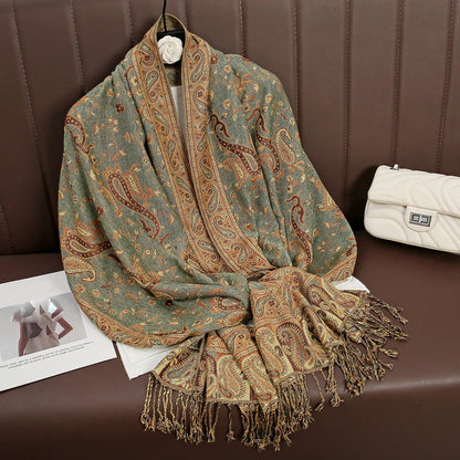 Green and gold paisley pashmina shawl with fringe on brown sofa, near white quilted purse
