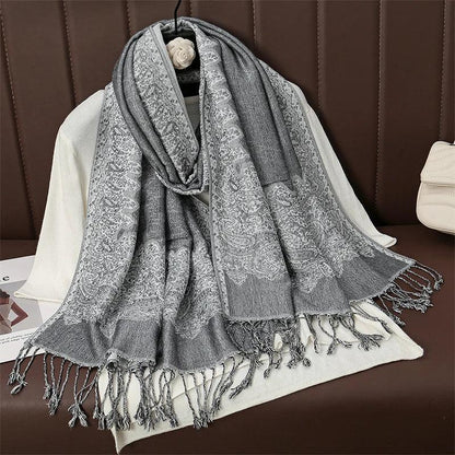 Gray paisley pattern scarf with fringe on white sweater, displayed on brown sofa