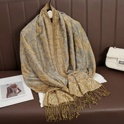 Gold and grey paisley pashmina scarf with fringe on brown bench, next to white purse