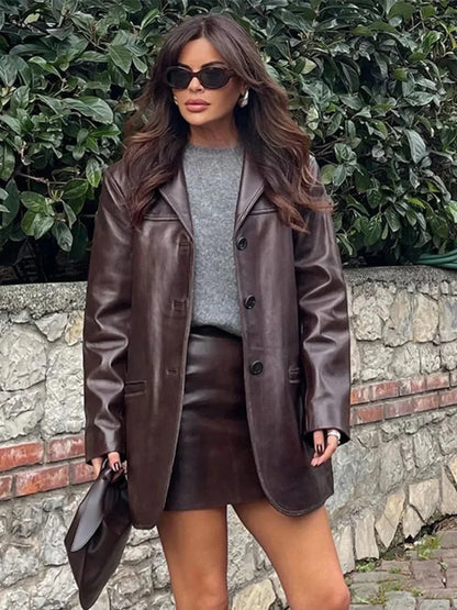 Woman wearing brown leather blazer and skirt with gray sweater, holding matching clutch outdoors