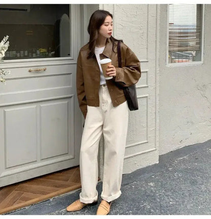 Woman wearing brown streetwear faux leather shirt jacket, white top, cream pants, holding coffee cup outside doorway