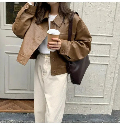 Woman wearing brown faux leather streetwear jacket holding coffee cup, paired with cream pants and brown shoulder bag