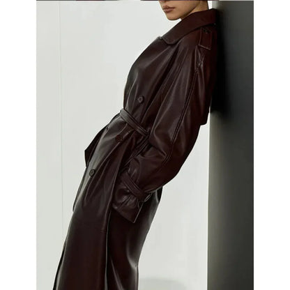Left side view of a model wearing a dark brown long faux leather trench coat with belted waist and button details