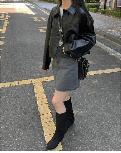 Woman wearing black faux leather streetwear shirt jacket with gray denim skirt and black boots on urban street