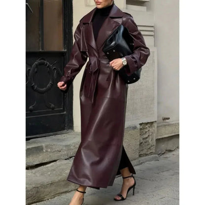 Side view of a woman wearing long dark brown faux leather trench coat with belt, black turtleneck, black heels, and holding a black bag
