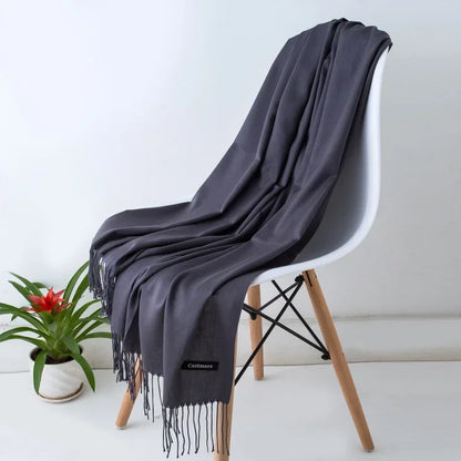 Dark gray cashmere scarf with fringes draped over a modern white chair with wooden legs, indoor plant nearby