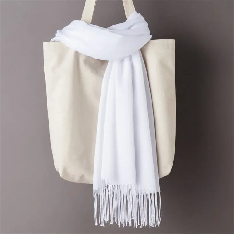 White cashmere scarf with fringe detail draped over beige tote bag against gray background