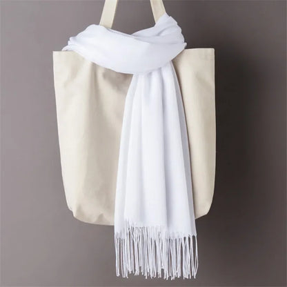 White cashmere scarf with fringe detail draped over beige tote bag against gray background