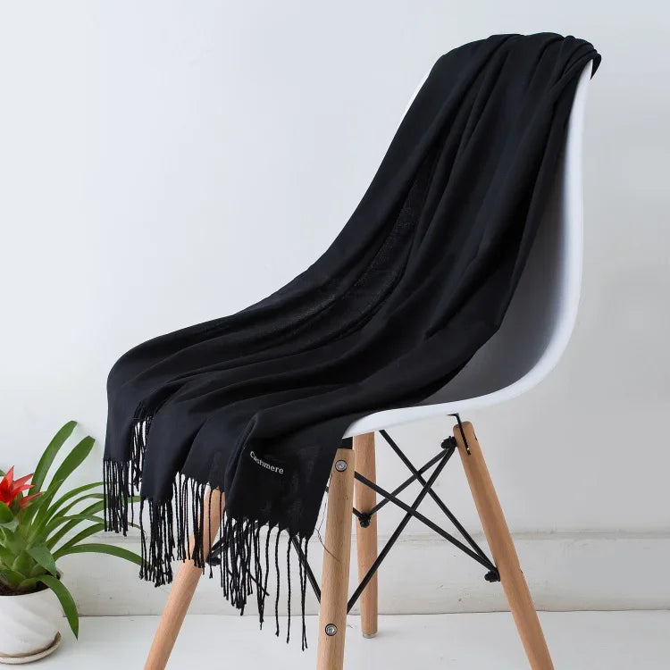 Black cashmere women's scarf with fringed edges draped over modern white chair with wooden legs in minimal setting