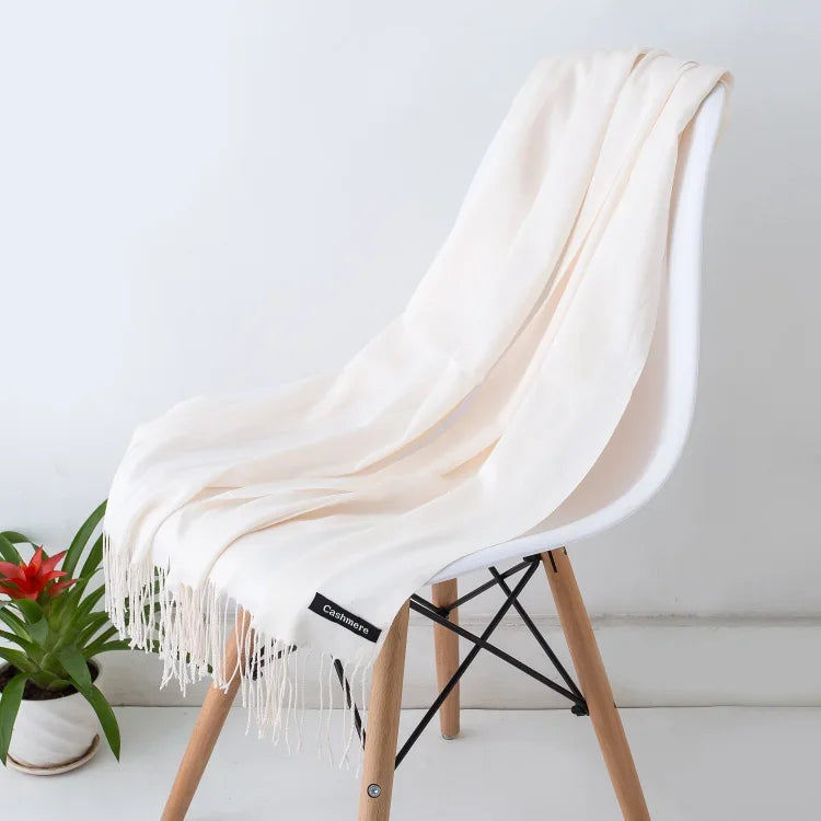 Soft ivory cashmere scarf with fringes draped over modern wooden chair beside green potted plant