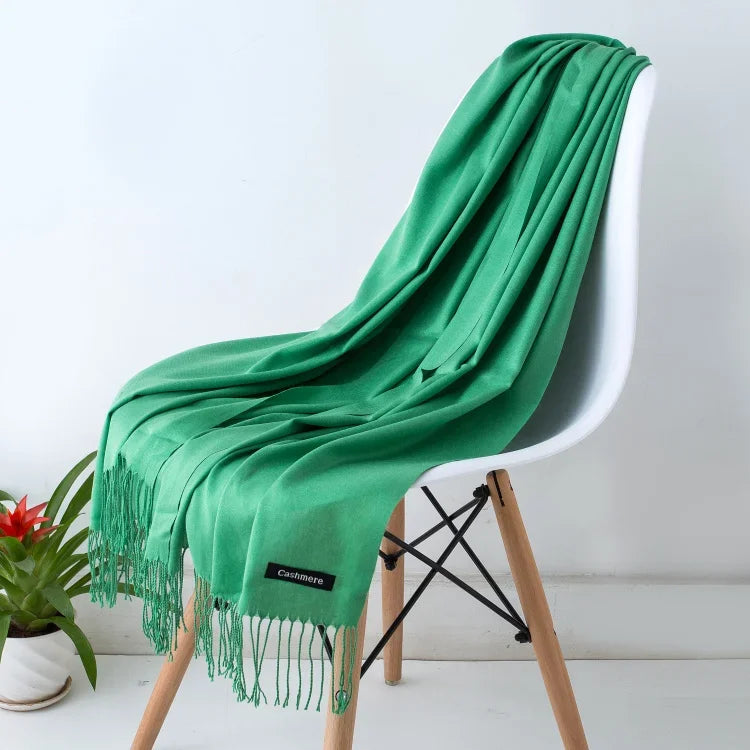 Green women's cashmere scarf draped over white modern chair with wooden legs, decorative plant nearby