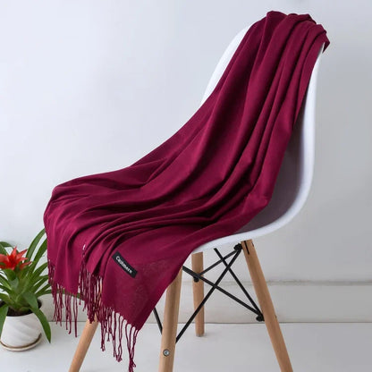 Luxurious solid color women's cashmere scarf in deep burgundy draped over modern white chair with wooden legs, cozy autumn winter shawl