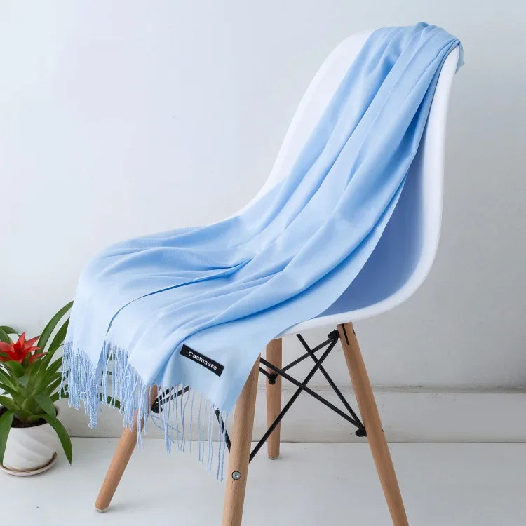 Light blue solid color cashmere scarf with fringes draped over a modern white chair with wooden legs in a minimalist setting