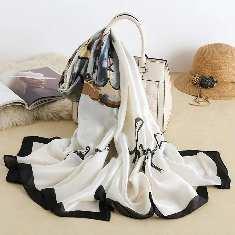 Elegant silk scarf with abstract print draped on a cream handbag, next to a straw hat