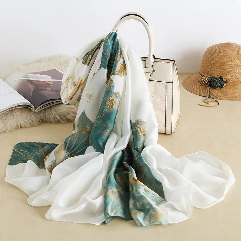 White and green floral silk scarf draped over a cream handbag, with straw hat and magazine nearby