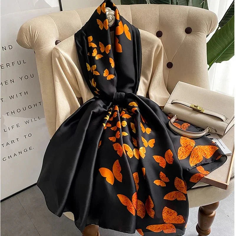 Black silk scarf with orange butterfly print draped on beige chair near cream handbag